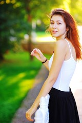 Wonderful portrait of a young smiling woman with beautiful brown hair wearing a white shirt and black skirt, standing in the rays of setting sun in summer Park