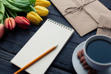 Notepad with pencil next to the tulips, coffee, and envelopes on wooden background. View from above...
