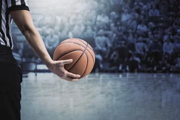 Poster basketball referee holding a basketball at a game in a crowded sports arena. Holding the ball in his hand during a timeout. Selective focus on the ball. The fans and crowd and basketball court © Brocreative