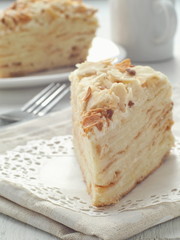 Piece of multi layered cake close-up. Mille feuille dessert. Crumbs decorated torte on white doily upon wooden table. Food background, blur. Selective focus on the front.