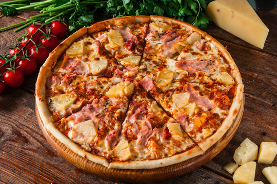 Delicious hawaiian pizza with ham and pineapple, served on rustic wooden background with cherry tomatoes, cheese and peppercorns. Italian restaurant menu photo.