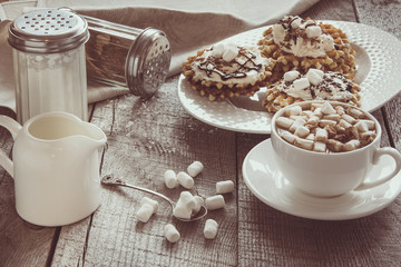 White cup of coffee with milk, marshmallow and homemade waffle with whipped cream with decor on top. Tasty breakfast. Vintage toned.