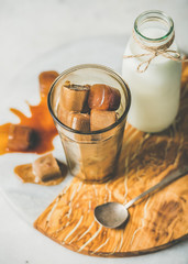 Glass with frozen coffee ice cubes and milk in bottle on olive wood and marble serving board over grey table background, selective focus