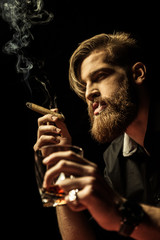 Handsome bearded man holding glass of whisky and smoking cigar on black