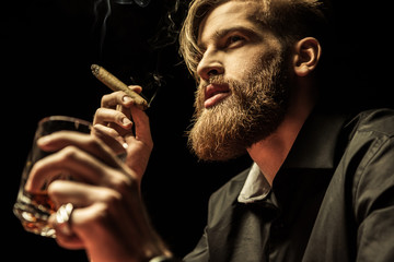 Handsome bearded man holding glass of whisky and smoking cigar on black