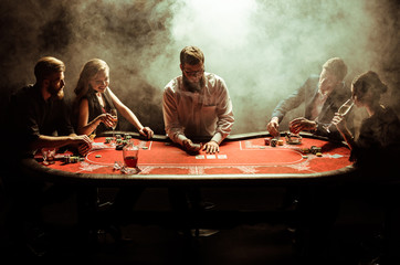 Elegant young men and women playing poker at table in smoke