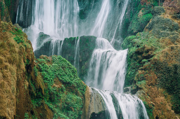 Beautiful photo of Ouzoud waterfall in Morocco with soft flowing water and large colored rocks. Green wild jungles on background