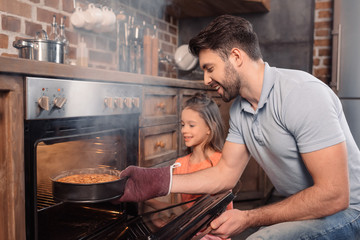 'Side view of smiling father and daughter taking cake from oven