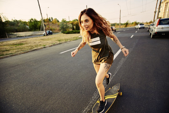 Beautiful young girl with tattoos riding on his longboard on the road in the city in sunny weather. Extreme sports