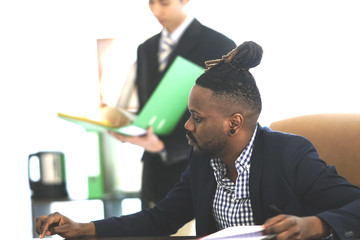 African American office worker with collected rasta hair at work. On background, Asian colleague...