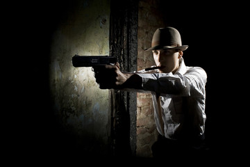 Portrait of a gangster dressed in retro suit with hat, smoking cigar and holding gun in hands. Ruined place.