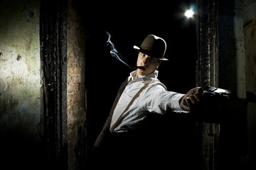 Portrait of a gangster dressed in retro suit with hat, smoking cigar and holding gun in hands. Ruined place.
