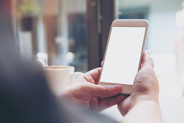 Mockup image of hands holding white mobile phone with blank white screen and hot coffee cup in loft cafe