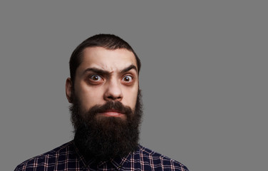 Surprised bearded man with wide open eyes and big moustache. Studio close up portrait of tanned skin hipster isolated on grey background. Serious and brutal person look to the camera. 