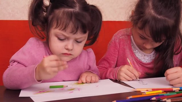 Children paint pencils. Two little girls draw on paper with crayons. Two sisters at home, on an orange couch draw on the table.