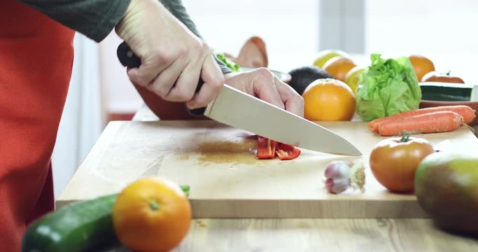 side view of hands of man cutting red pepper slices on brown wood plank with red and orange apron and green shirt
