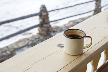 Enamel mug with strong tea and tea bag on a wooden fence on a snowy winter background.