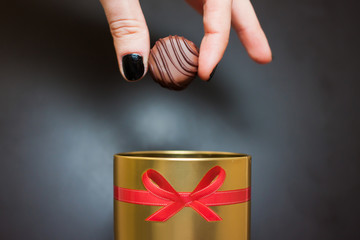 mano che tiene caramella di cioccolato della donna, tirato fuori da una scatola d'oro rotonda con un fiocco rosso su sfondo nero