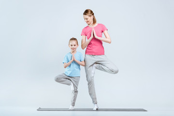 Fototapeta na wymiar Smiling sporty mother and daughter practicing yoga together, Vrikshasana yoga pose or tree pose on white