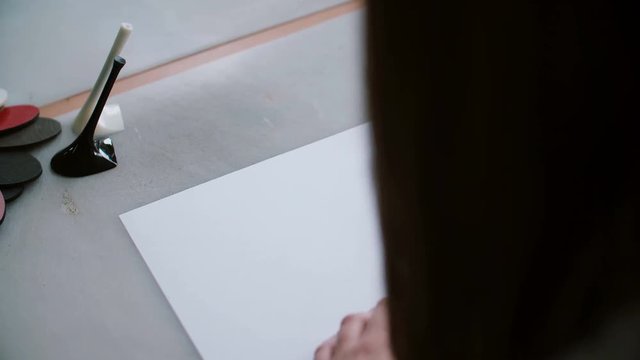 Back view of woman sitting at table with heel and textile samples on it, drawing sketch on paper. Slider left. 4K