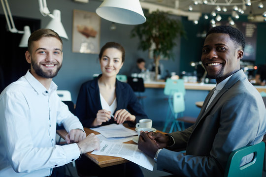 Portrait of three confident business people sitting at table during meeting in cafe, one of them African, all smiling looking happily at camera