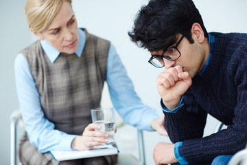 Portrait of young depressed man wearing sweater and glasses opening up to his psychologist about mental problems