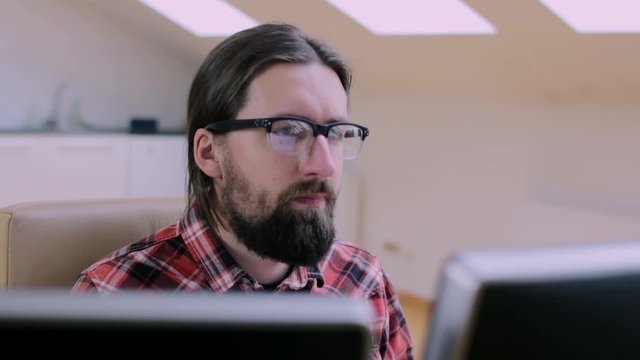 Bearded man in glasses working in office with 2 monitors