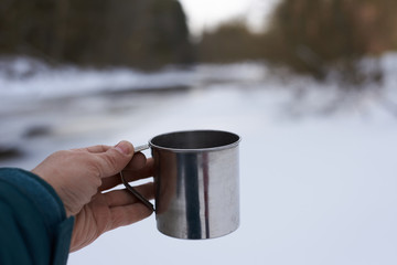 Tourist hand with metal cup of tea or coffee outdoor in cloudy day with frozen river background. Hiking break time. Eye view.