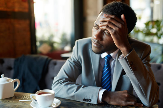 Portrait Of Handsome African American Man Wearing Business Suit Looking Away To Window, Pensively  Resting Head On Hand While Sitting At Table In Modern Restaurant During Coffee Break