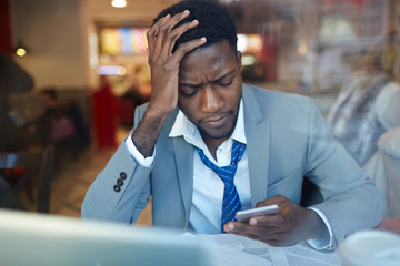 Obraz premium Businessman looking stressed and resting his head on hand while typing message in smartphone sitting at counter in coffee shop, shot behind glass window