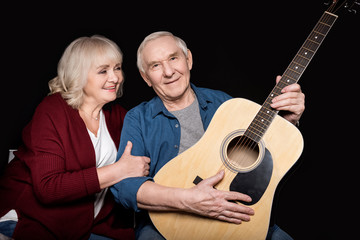 portrait of senior man holding guitar with wife near by on black