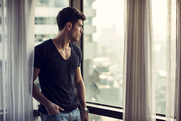 Sexy handsome young man standing next to window curtains, looking out