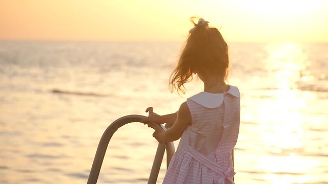 beautiful little girl standing on the pier holding railing and looks at the setting sun, then looks down into the water