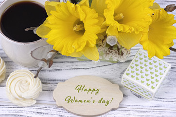 Cup of coffee with marshmallow, green gift box, yellow flowers on white wooden background and lettering happy women's day english.