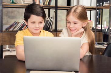 Cute smiling boy and girl sitting at table and using laptop together