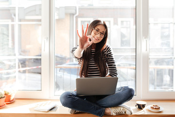 Asian woman on windowsill with laptop showing ok sign