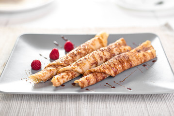 Three Russian thin pancakes rolled into a tube with raspberries and caramel sauce on gray plate