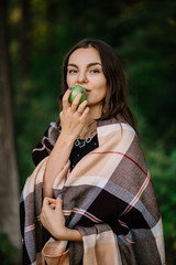 Fototapeta premium beautiful brunette girl were wrapped in plaid blanket in nature is eating a green Apple looking down