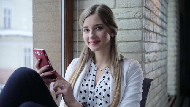 Young beautiful blonde start-upper sits in the modern office, texting on her mobile phone then looks to the camera and smiles.