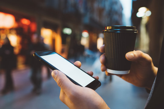 Close-up image of male hands holding coffee to go and using modern smartphone with blank screen with area for your design or logo, night city street in the background