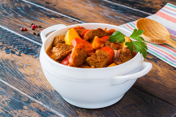 Traditional Hungarian dish, goulash, slow-cooked with beef, potatoes, carrot, tomatoes, paprika, onion and bell peppers. White bowl on wooden table.