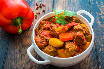 Traditional Hungarian dish, goulash, slow-cooked with beef, potatoes, carrot, tomatoes, paprika, onion and bell peppers. White bowl on wooden table.