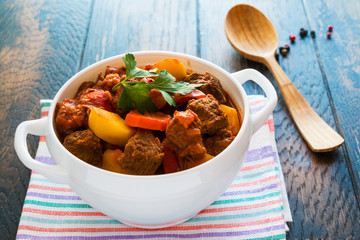 Traditional Hungarian dish, goulash, slow-cooked with beef, potatoes, carrot, tomatoes, paprika, onion and bell peppers. White bowl on wooden table.