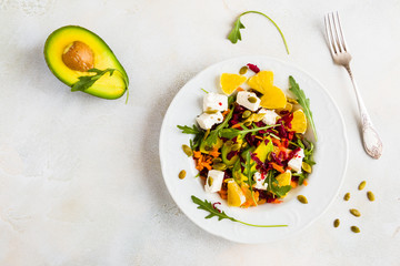 Healthy vegetarian salad with raw beetroot, carrot, arugula, avocado, orange, cheese and pumpkin seeds on white plate, top view.