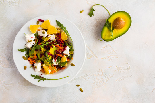 Healthy Vegetarian Salad With Raw Beetroot, Carrot, Arugula, Avocado, Orange, Cheese And Pumpkin Seeds On White Plate, Top View.