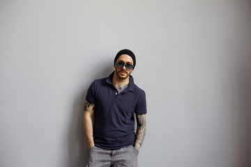 Obraz premium A stylish young Caucasian man wearing sunglasses with a beard and tattoos looking at camera with serious face expression, looks self-confident and calm. Masculinity and style concept