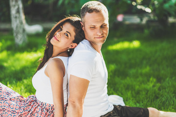 Fototapeta premium Cute couple in love sitting on a picnic blanket outdoor. Caucasian happy dreamy man and smiling brunette woman sitting on green grass in park.