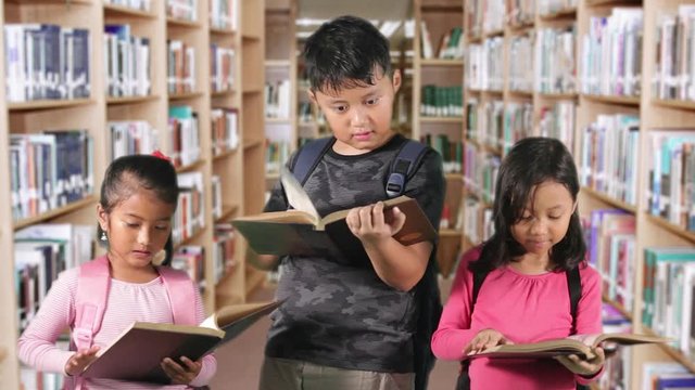 Three elementary school students standing in the library while reading books