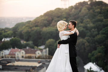 Sensual beautiful bride and handsome groom hugging tenderly on a hilltop overlooking the panorama of the city on sunset