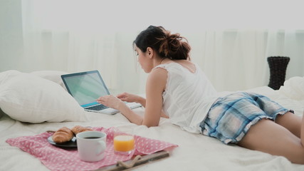 Obraz na płótnie Canvas Young woman using laptop during breakfast lying on white bed at home. Brunette girl typing on pc computer.